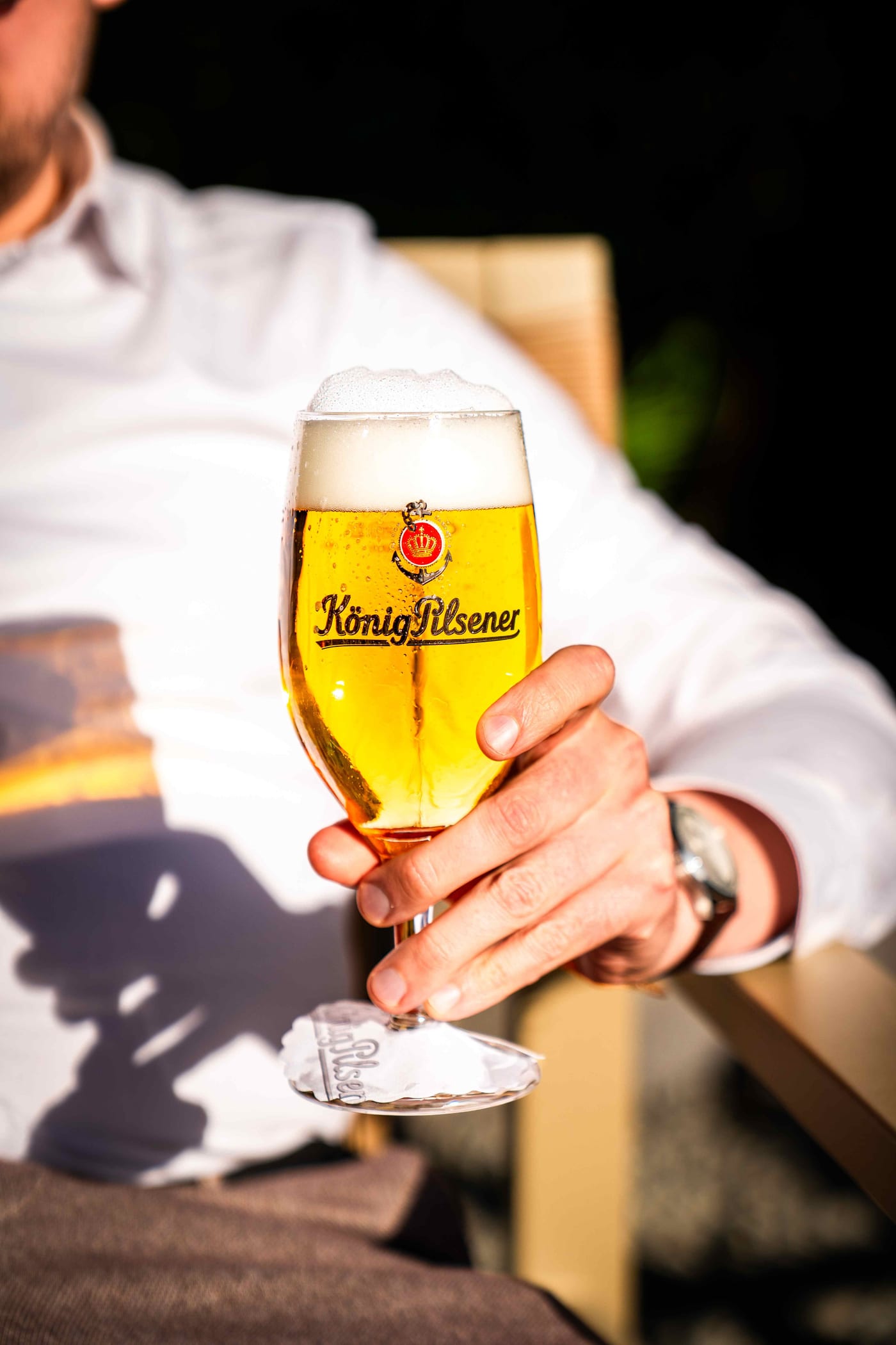 Hand hält ein frisch gezapftes König Pilsener im Glas im Licht der Abendsonne