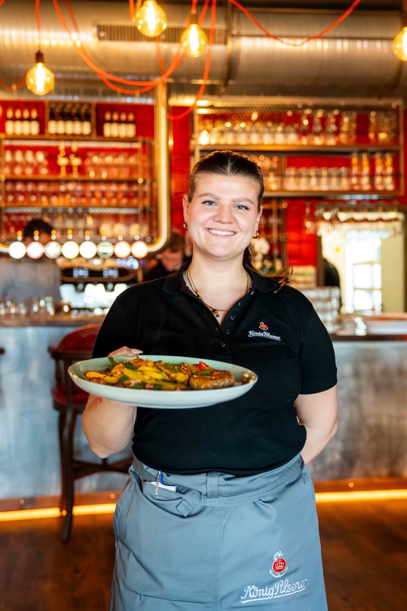 Lächelnde Servicekraft im König-Pilsener-Polo serviert einen Teller im Wirtshaus Moers