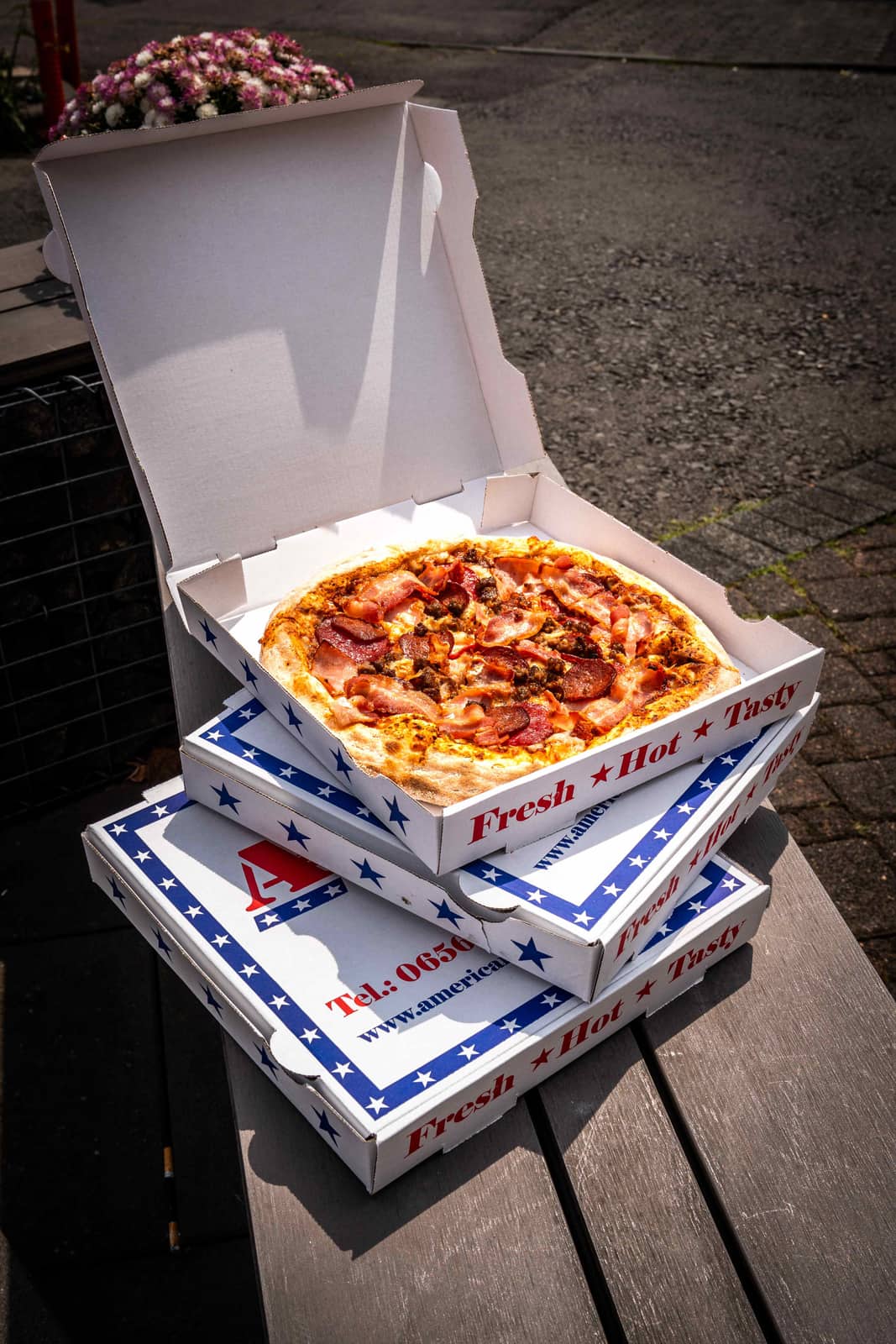 Frisch belegte American Pizza mit Speck in gestapelten Pizzakartons im Sonnenlicht