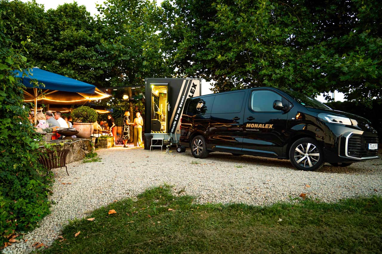Schwarzes Monalex Event-Fahrzeug mit mobiler Bühne bei einem Gartenevent in der Abenddämmerung