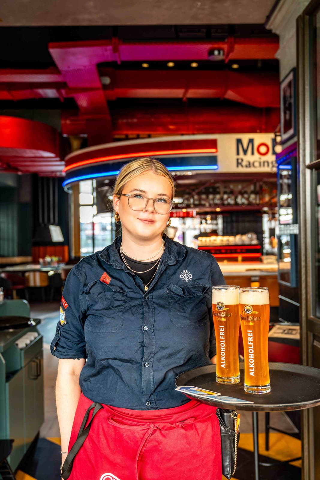 Servicekraft im MoT Racing Restaurant serviert zwei Gläser alkoholfreies Weissbier auf einem Tablett